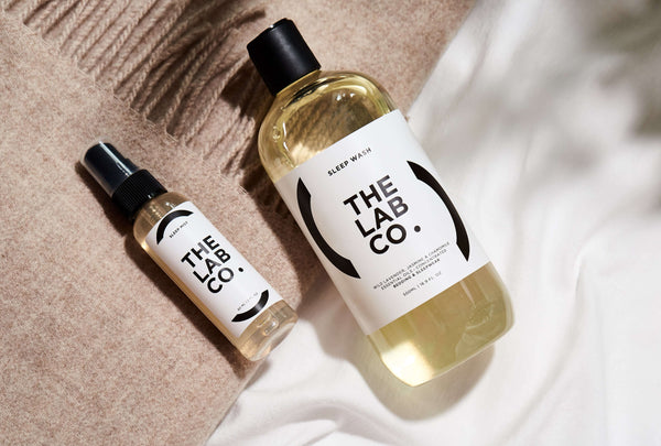 The Lab Co. Sleep Laundry Detergent and Fabric Mist placed on top of white bedsheets and a pink throw